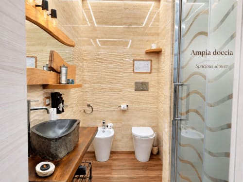 Baño moderno con lavabo de piedra negra, bidet blanco, tocador de madera y paredes con azulejos beige texturados en la habitación Basileus del B&B I