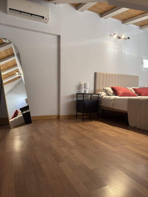 Dormitorio luminoso en apartamento madrileño con vigas de madera, cama con cabecero beige, almohadas rojas, mesita negra y espejo de cuerpo entero