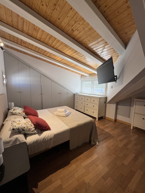 Moderno dormitorio abuhardillado en apartamento madrileño con vigas de madera, cama queen, almacenamiento y televisor