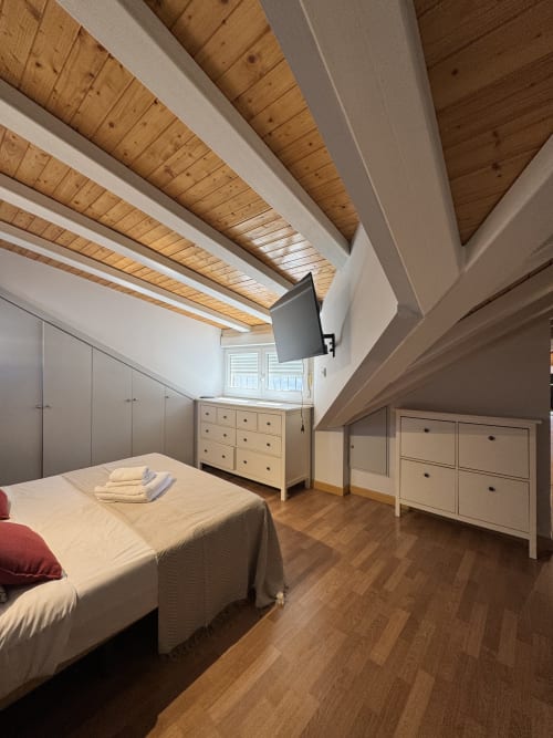 Moderno dormitorio en ático madrileño con vigas de madera vista, paredes blancas, cama y muebles de almacenaje junto a la ventana