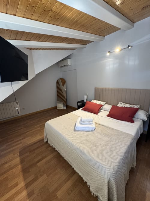 Moderno loft dormitorio en Madrid con vigas de madera, cabecero beige, almohadas rojas y suelos de madera