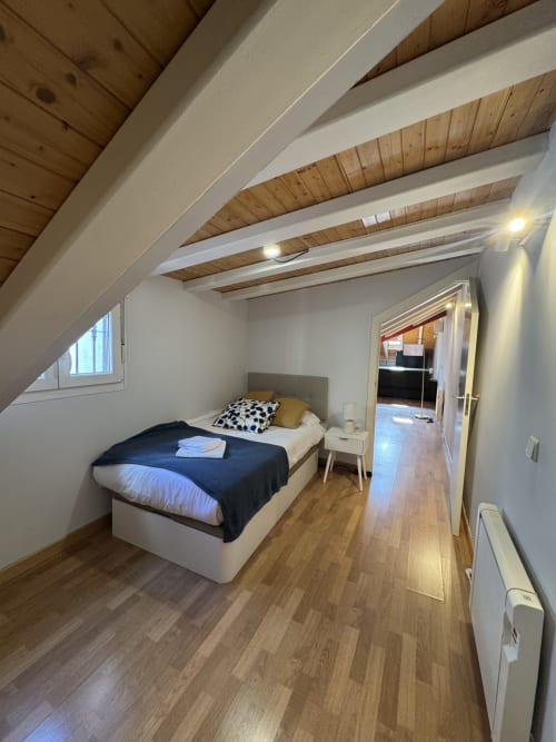 Luminoso dormitorio en buhardilla madrileña con vigas de madera vista, cama, claraboya y suelo de madera