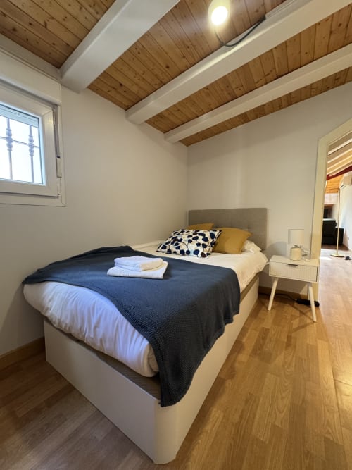 Dormitorio luminoso en apartamento madrileño con techo de vigas de madera, cama moderna con manta azul, almohada amarilla y vistas a la ciudad desde