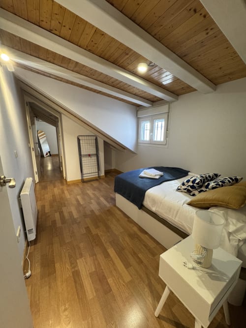 Luminoso dormitorio abuhardillado en la zona de Colón de Madrid con vigas de madera, suelos de parquet y vistas de la ciudad