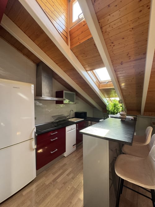 Cocina moderna en ático con tragaluz en apartamento madrileño, armarios rojos, encimera negra y vigas de madera