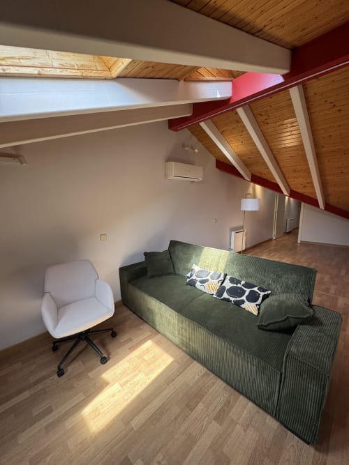 Salón de ático moderno en Madrid con sofá verde, silla blanca, vigas de madera y vistas de la ciudad desde la calle Colón