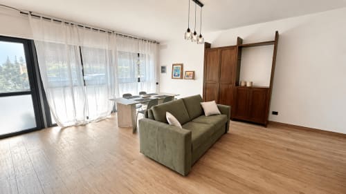 Soggiorno moderno con divano verde oliva, pavimentazione in legno, tavolo da pranzo e vista sulla città all'appartamento Tolstoi 15 a Saronno