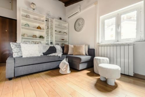 Soggiorno moderno della Luxury Trevi House Roma con divano grigio, scaffali bianchi, pavimento in legno e vista sulla città