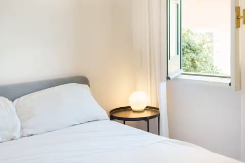 Camera moderna al B&B Oliveri 25 di Procida con biancheria bianca, testata grigia, tavolino rotondo con lampada luminosa e finestra aperta