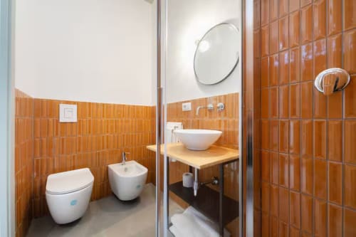 Bagno moderno con piastrelle arancioni, lavabo a coppa bianco, WC, bidet e specchio circolare presso B&B Oliveri 25 a Procida