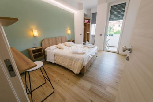 Camera moderna a Camera Olivo a Verona con parete accent verde salvia, letto color crema, scrivania in legno e porta in vetro che conduce a