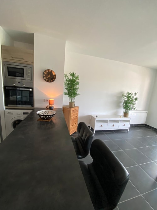 Appartement moderne à La Rochelle avec cuisine au plan de travail sombre, chaises noires, plantes en pots, horloge murale et mobilier contemporain