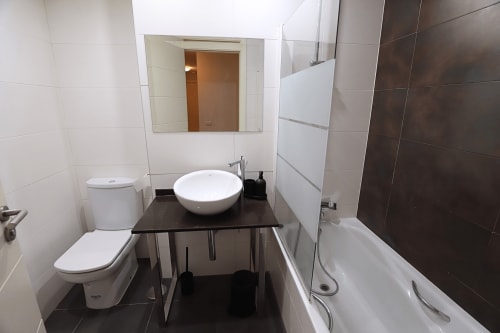Baño moderno en apartamento de Madrid Río con lavabo redondo blanco, inodoro, bañera y detalles en azulejos oscuros