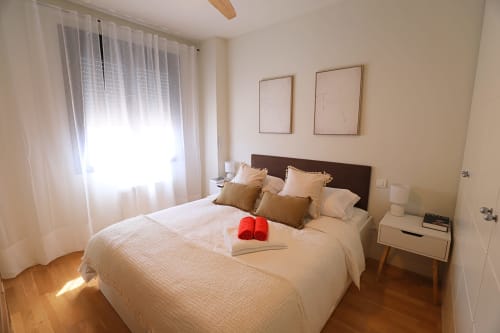 Dormitorio moderno en apartamento de Madrid Río con cama matrimonial, cabecero marrón, almohadas doradas y pantuflas rojas sobre ropa de cama blanca