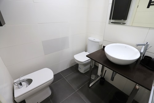 Baño moderno en apartamento de Madrid Río con dos lavabos, inodoro, azulejos blancos y piso negro