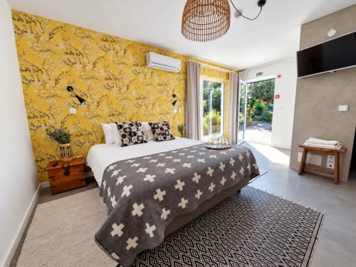 Modern bedroom with bold yellow floral wallpaper, white bedding, dark patterned throw, wooden nightstand, and garden views at