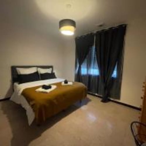 Chambre moderne dans un appartement de Perpignan avec lit, plaid jaune, rideaux noirs, plafonnier et balcon avec vue sur la ville