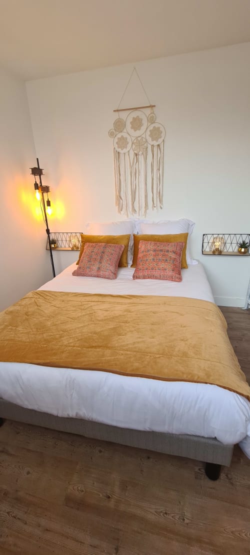 Chambre moderne aux Suites Du Port, Arcachon avec couette dorée, coussins corail, macramé mural et luminosité douce