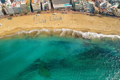 Vista aérea de la playa de Arucas con olas turquesas, costa arenosa, bañistas y coloridos edificios frente al mar