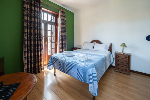 Quarto luminoso na pousada Casa Augusta em Penafiel com roupa de cama azul, móveis de madeira, parede de destaque verde e porta de vidro