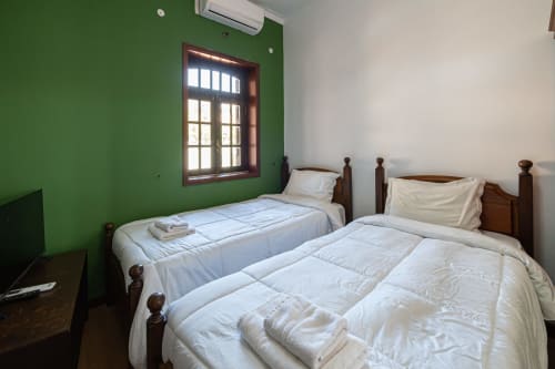 Quarto iluminado da pousada Casa Augusta com duas camas simples, paredes verdes, estrutura de madeira, ar condicionado e janela com vista para a montanha