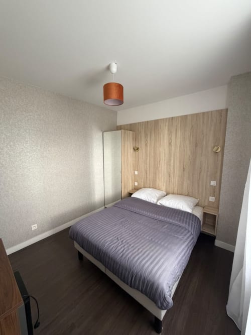 Chambre moderne à l'Hôtel le Dellys à Colombes avec tête de lit en bois, literie grise, luminaire suspendu et sol foncé