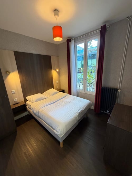 Chambre moderne à l'Hôtel le Dellys à Colombes : lit queen-size, luminaire rouge pendant, portes-fenêtres avec vue sur la ville