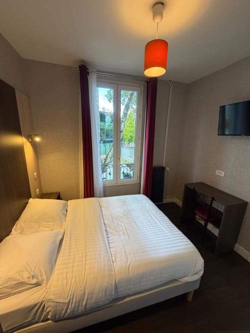 Chambre moderne à l'Hôtel le Dellys à Colombes avec lit queen, télévision et portes-fenêtres donnant sur la région