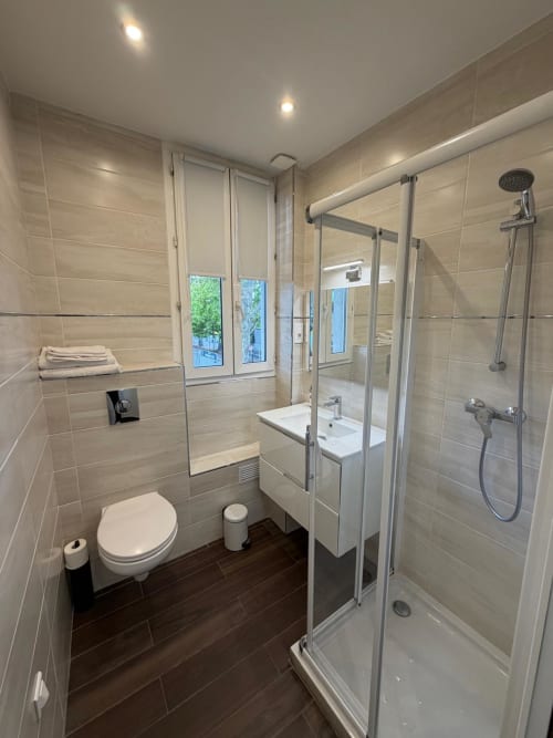 Salle de bain moderne de l'Hôtel le Dellys Colombes avec douche vitrée, lavabo blanc, toilettes et carrelage beige