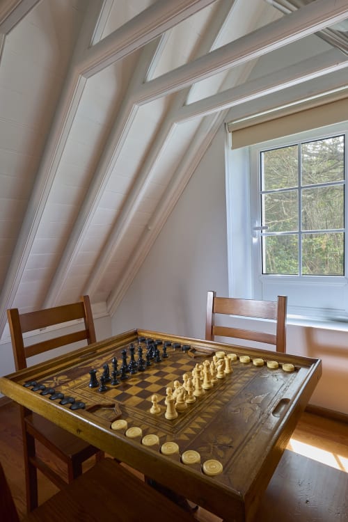 Quarto no sótão com mesa de jogos de xadrez e damas, cadeiras de madeira, teto inclinado e janela com vista para árvores em Casa Dos