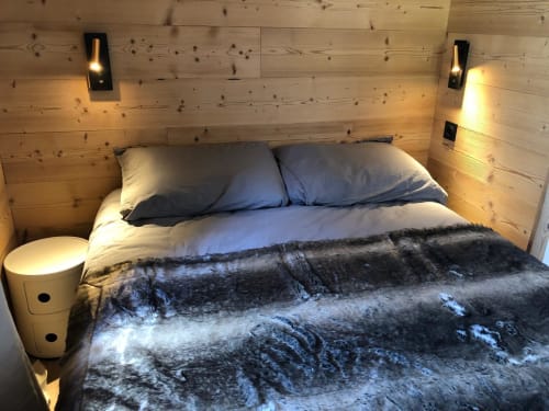 Chambre douillette aux boiseries, lit double, jeté de fourrure sombre, oreillers, lampe de chevet et meuble de rangement blanc à Montgenèvre