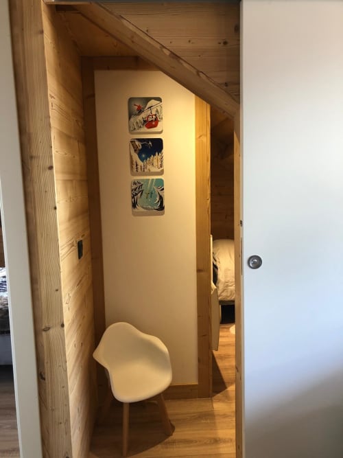 Couloir étroit dans l'appartement de Montgenèvre avec chaise blanche, poutres en bois et trois œuvres d'art sur le ski encadrées sur mur crème