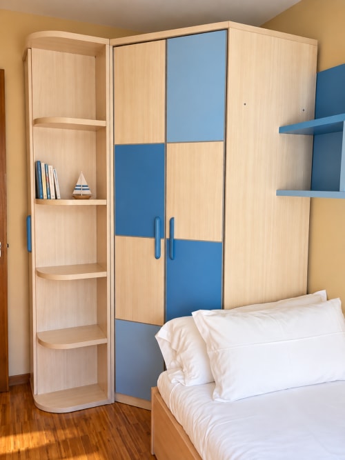 Dormitorio moderno con armario de madera clara, puertas correderas azules, estantería abierta, almohadas blancas y piso de madera en Aire