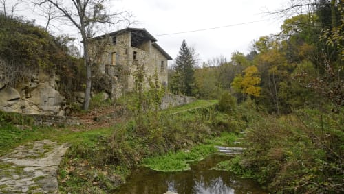 Edifici de pedra històric a l'Hostal Collsacabra de L'Esquirol, envoltat de bosc frondós i un petit rierol