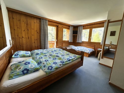 Spacieuse chambre triple à l'Hôtel les Bouquetins à Zinal avec murs en bois, deux lits, vue sur balcon et décor alpin.