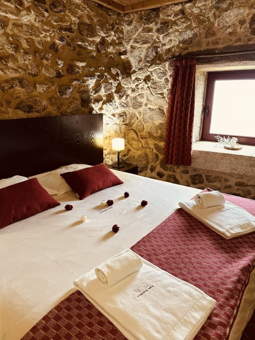 Rustic stone bedroom at Aconchego Rural with white bedding, burgundy pillows, wooden headboard, and ambient lighting