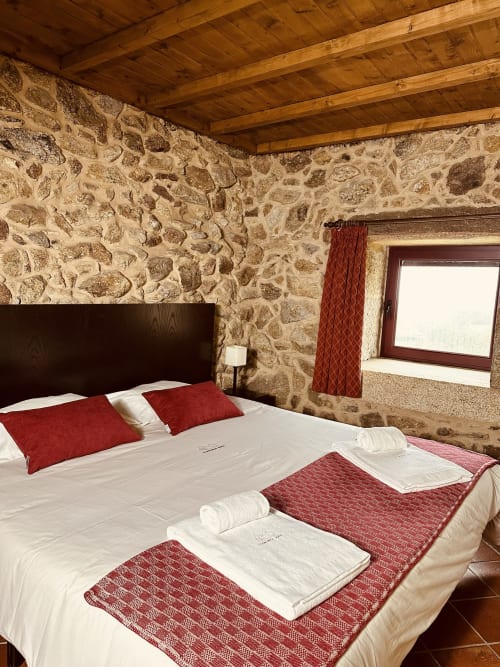 Rustic stone cottage bedroom with wooden beams, dark headboard, red accents, and window with mountain views at Aconchego