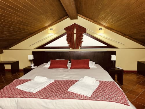 Rustic attic bedroom at Aconchego Rural cottage with wooden beams, queen bed, red pillows, and mountain views from skylights