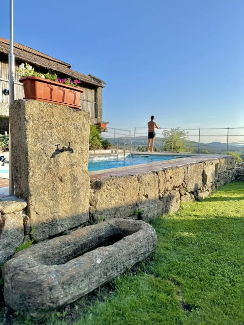 Shirtless man standing at poolside of private cottage with blue pool, stone walls, flowering planter, mountains beyond at