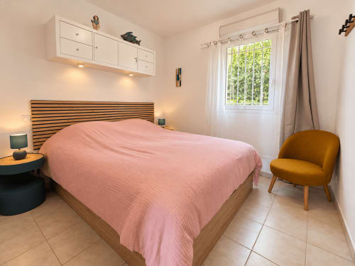Chambre lumineuse avec literie rose, tête de lit en lattes de bois et fauteuil ocre à l'appartement Calvi à L'Île-Rousse