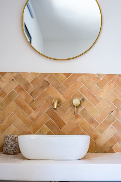 Lavabo blanc moderne avec robinetterie dorée et miroir encadré or au-dessus du dosseret en carreaux terre cuite à chevrons au Domaine Mas