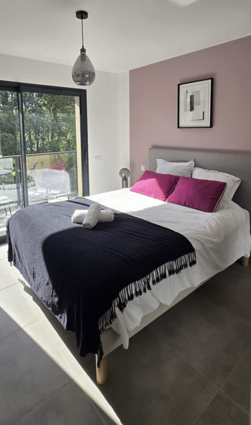 Chambre moderne avec mur accent rose, oreillers magenta, plaid noir et porte vitrée donnant sur piscine aux Villas d'A