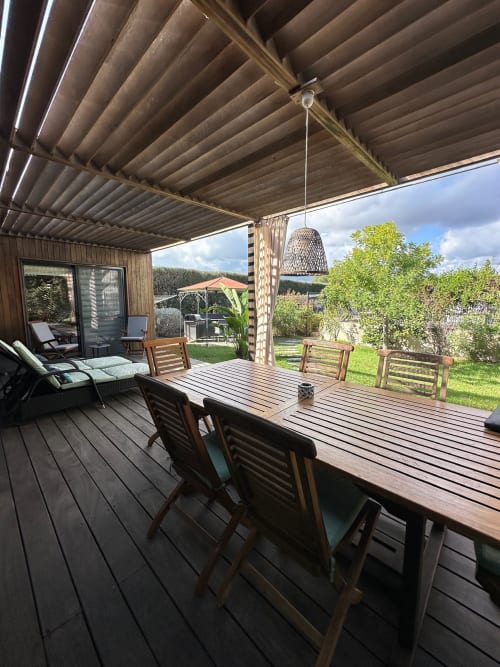 Terrasse couverte de la Villa Maé à Ghisonaccia avec table de salle à manger en bois, chaises et luminaire suspendu en osier dominant le jardin