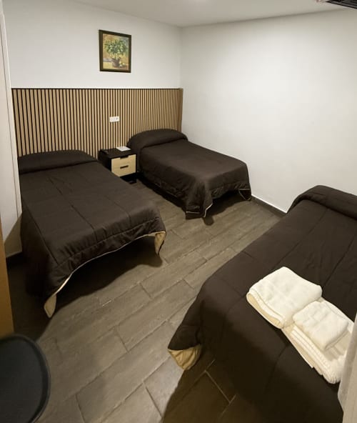 Habitación triple del Hostal3hermanos en Getafe con tres camas, ropa de cama oscura, cabecero de madera y toallas blancas