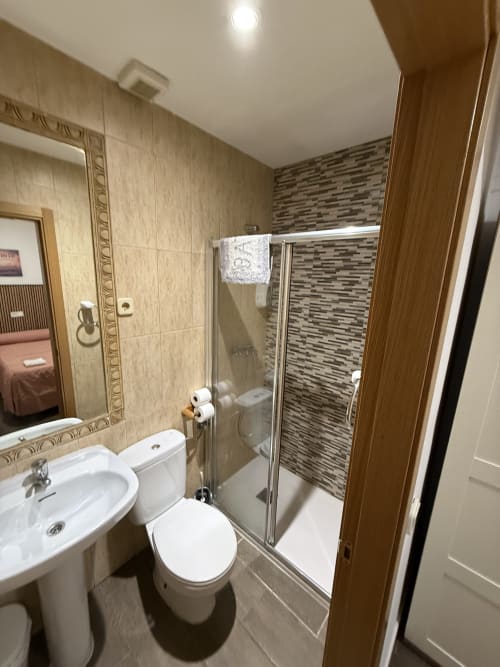 Baño moderno con cabina de ducha de vidrio, inodoro blanco, lavabo de pedestal y paredes alicatadas en Hostal3hermanos en Getafe