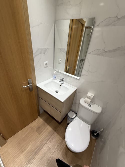 Cuarto de baño moderno en Hostal3hermanos Getafe con lavabo blanco, inodoro, paredes de mármol y piso de madera
