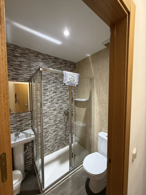 Baño moderno en Hostal3hermanos Getafe con ducha de azulejos rayados, inodoro blanco, lavabo y televisor empotrado