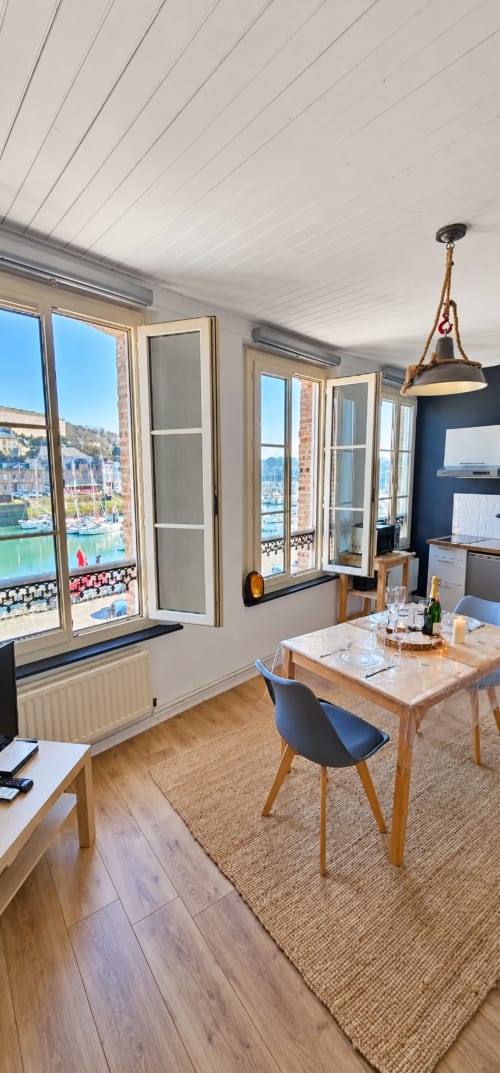 Salle à manger contemporaine avec vue sur le port de Saint Valery En Caux par de grandes fenêtres, table en bois, chaises bleues, blanc