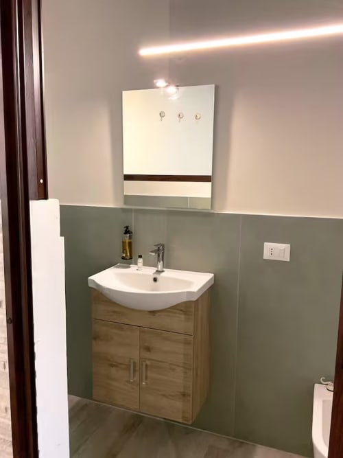 Salle de bain moderne au Sikania Agri Relais avec lavabo blanc incurvé, meuble en bois, carrelage vert et miroir illuminé