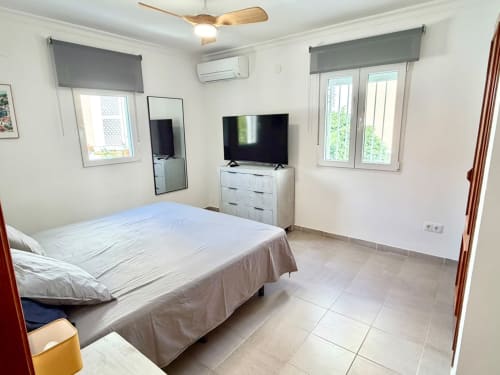 Dormitorio luminoso en casa rural Casa Mar Blau, Valencia, con cama, TV, cómoda blanca, ventanas con vistas al mar y comodidades modernas